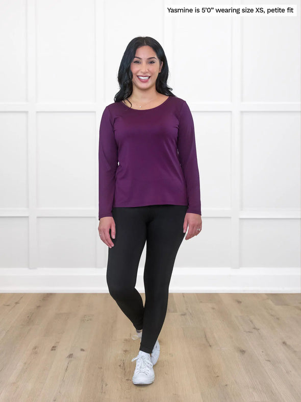 Miik model Yasmine (5'0", xsmall) smiling while standing in front of a white wall wearing Miik's Shannon reversible neckline long sleeve tee in port, petite fit, with a black legging #color_port