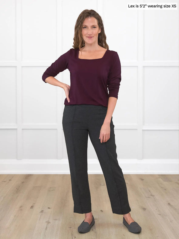 Miik model Lex (5'2", xsmall) smiling while standing in front of a white wall studio wearing a charcoal dress pant in petite fit along with Miik's Shyla reversible square neck top in port 
#color_port