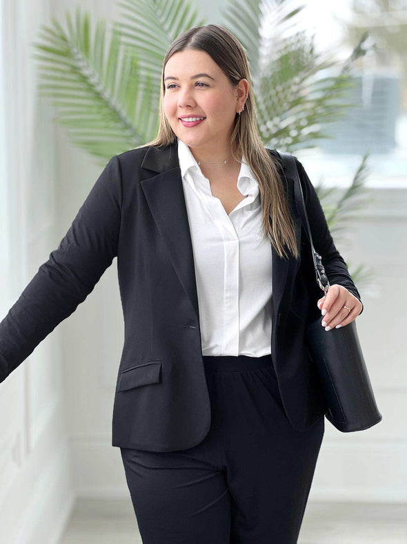 Miik model Christal (5'3", large) leaning against a window looking away wearing Miik's SIenna girlfriend blazer in black with a collared shirt in white and black pants  #color_black