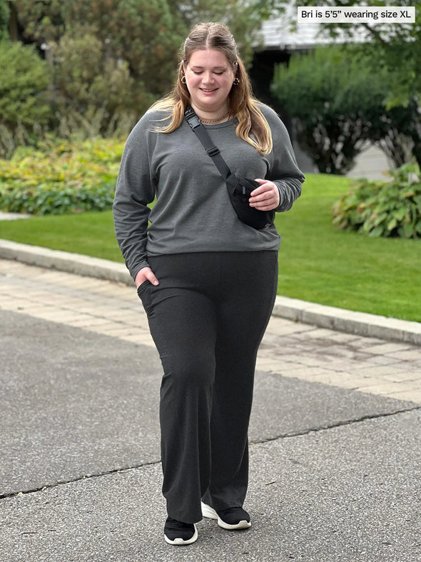 Miik model Bri (5'5", xlarge) smiling and looking down wearing Miik's Sierra high waisted pocket pant in charcoal with a sweatshirt in granite and a black belt bag and sneakers #color_charcoal