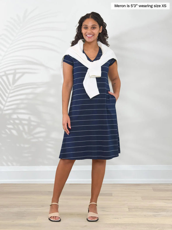 Miik model Meron (5'3", xsmall) smiling whie standing in front of a white wall wearing Miik's Silas t-shirt dress with pockets in navy pinstripe with a sweater tied over her shoulders #color_navy-pinstripe