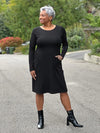 Miik model Keethai (5'5", medium) smiling and looking away wearing Miik's Soraya long sleeve scoop neck pocket dress in black with boots in the same matching colour 