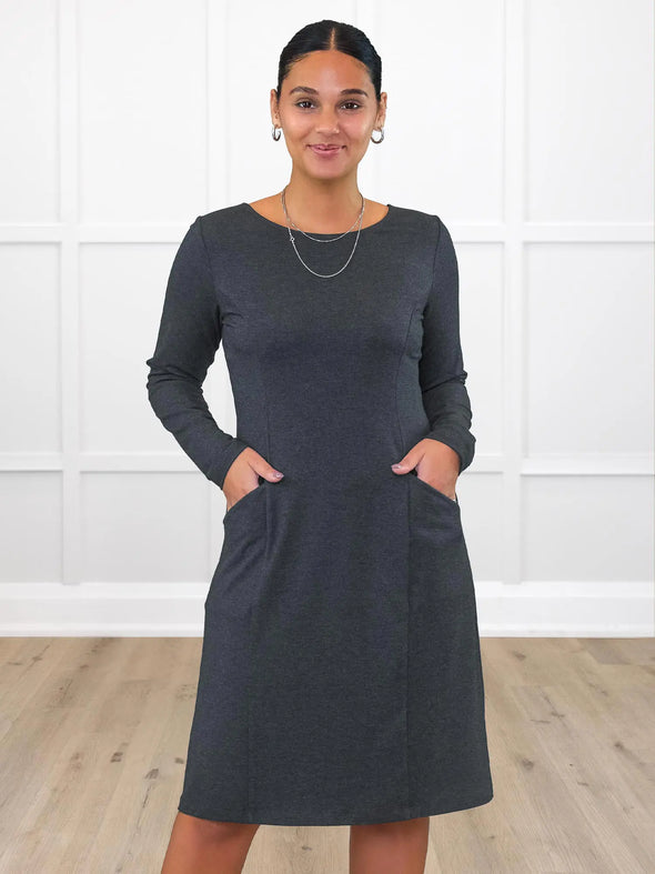 Miik model Meron (5'3", xsmall) smiling while standing in front of a white wall wearing Miik's Soraya long sleeve scoop neck pocket dress in charcoal #color_charcoal 