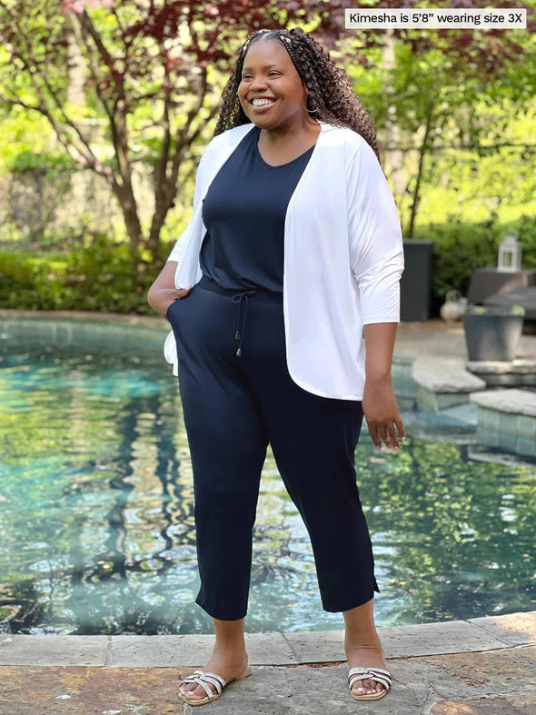 Miik model plus size Kimesha (5'8", 3x) smiling while standing in front of a pool wearing Miik's Stef v-neck open-back capri jumpsuit in navy with a cropped cardigan in white 
#color_navy