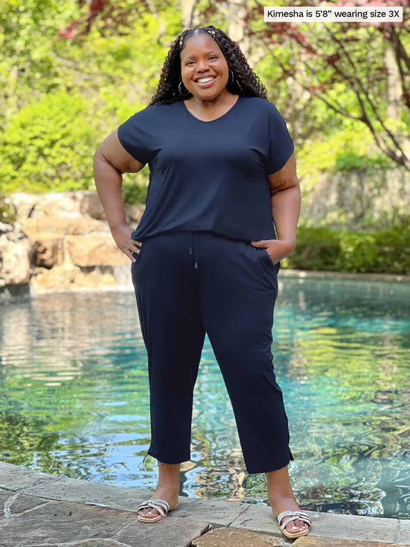 Miik model plus size Kimesha (5'8", 3x) smiling while standing in front of a pool wearing Miik's Stef v-neck open-back capri jumpsuit in navy #color_navy