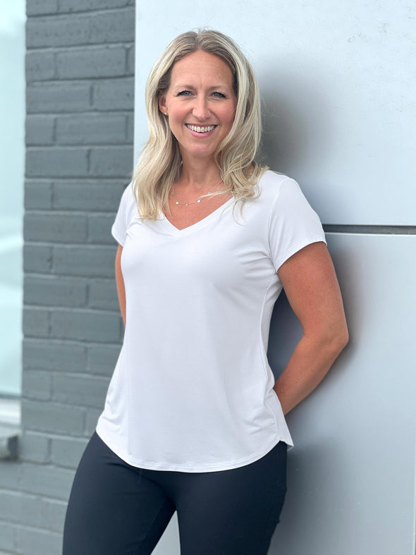 Woman leaning against a grey wall, smiling while wearing Miik's Sutton v-neck classic tee in white with black pants #color_white