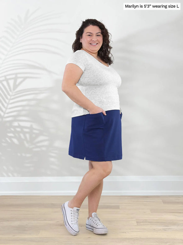 Miik model Marilyn (5'3", large) smiling while standing sideway in front of a white wall wearing Miik's Tessa high waisted pocket skort in marine blue with a pearl melange basic tee #color_marine-blue