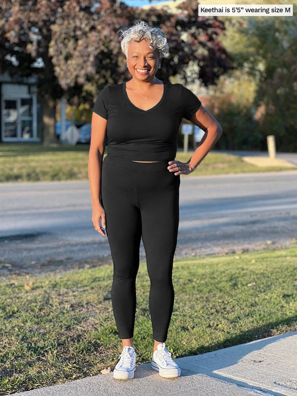 Miik model Keethai (5'5", medium) smiling wearing Miik's Tinsley mid-rise fleece legging in black with a v-neck tee in the same colour and white sneakers #color_black 