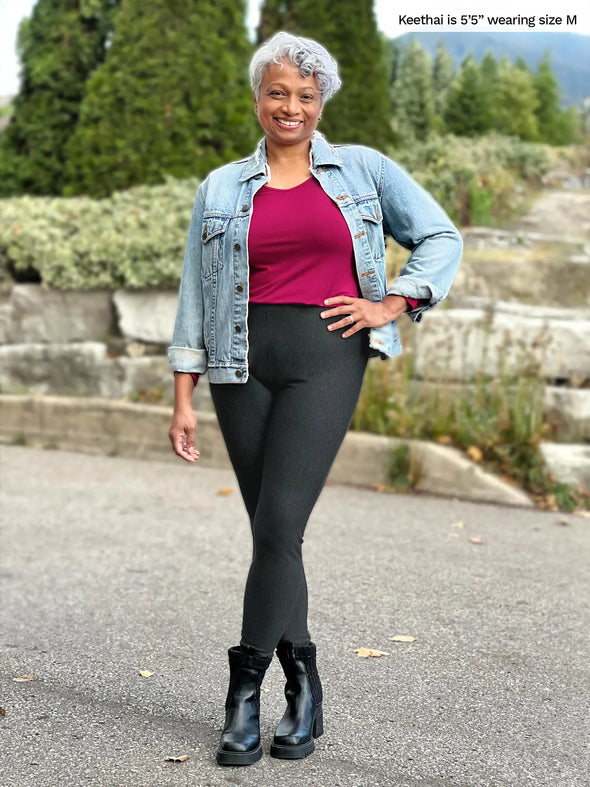 Miik model Keethai (5'5", medium) smiling wearing Miik's Tinsley mid-rise fleece legging in charcoal with a cerise v-neck top, a denim jacket and black boots #color_charcoal 