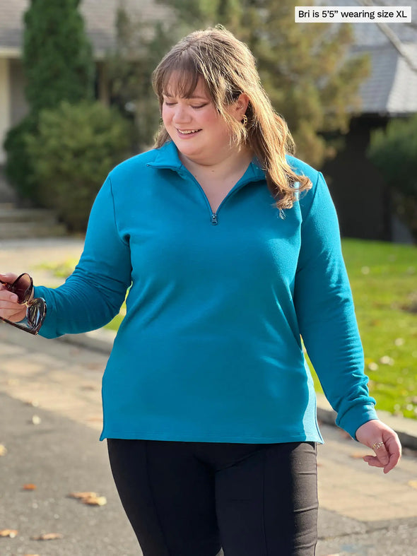 Miik model Bri (5'5", xlarge) smiling and looking down wearing a black pant along with Miik's Tristan 1/4 zip fleece pullover in teal melange #color_teal-melange 