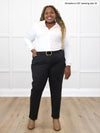 Miik model Kimesha (5'8", 3x) smiling while standing in front of a white wall wearing Miik's Uma mid-rise straight leg ponte pant in black, with a black belt and a white collared shirt #color_black