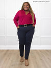 Miik model Kimesha (5'8", 3x) smiling while standing in front of a white wall wearing Miik's Uma mid-rise straight leg ponte pant in navy melange, with a black belt and a cerise collared shirt #color_navy-melange