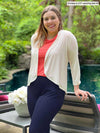 Miik model Christal (5'3", medium) smiling while leaning against to a outdoor chair wearing a navy pant, papaya melange tee and Miik's Wesley cropped cardigan in natural 
