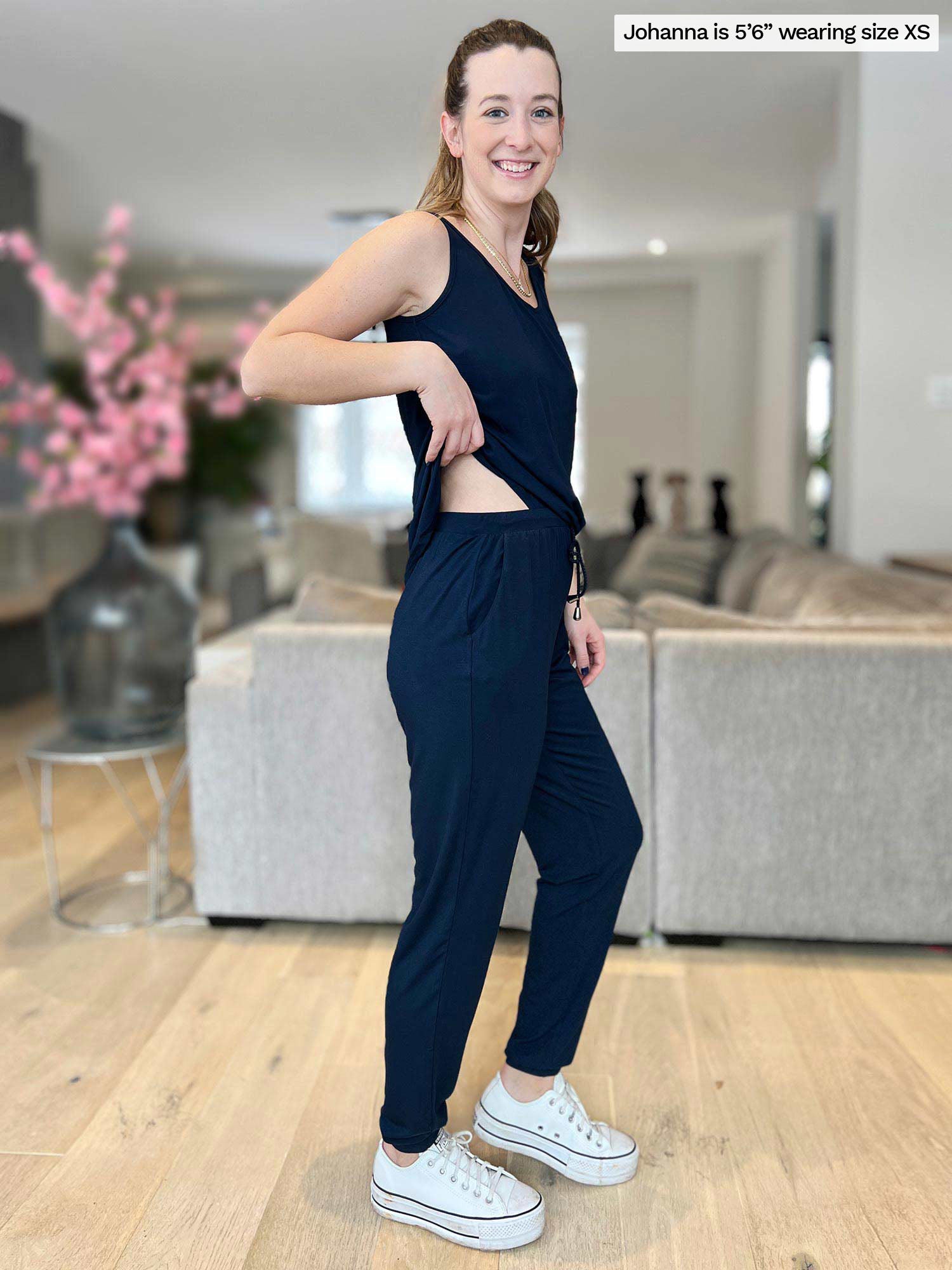 Miik model Johanna (5 foot 6, size extra small) wearing the Perle open-back tank top jumpsuit in a living room, smiling while lifting the back of the jumpsuit&